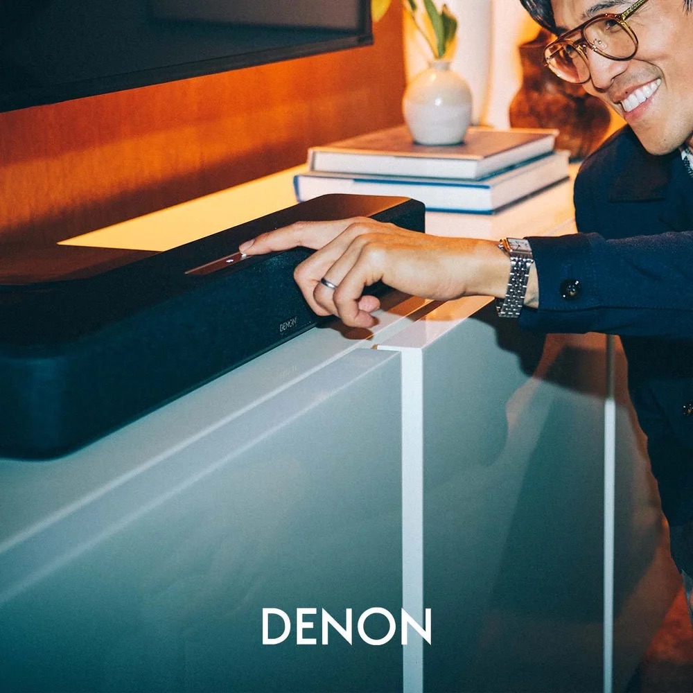 Denon Home Sound Bar below a wall-mounted TV in a warm, wood-panelled living room; a smiling man in glasses and a navy coat leans in to press a control on the bar.